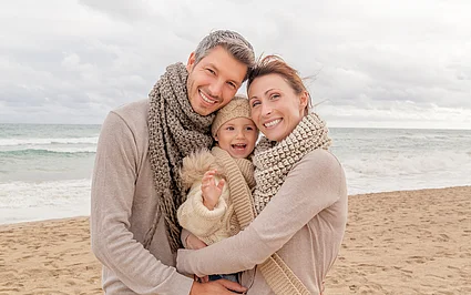 Familie mit kleinem Kind umarmt sich lächelnd an einem windigen Strand vor dem Meer.
